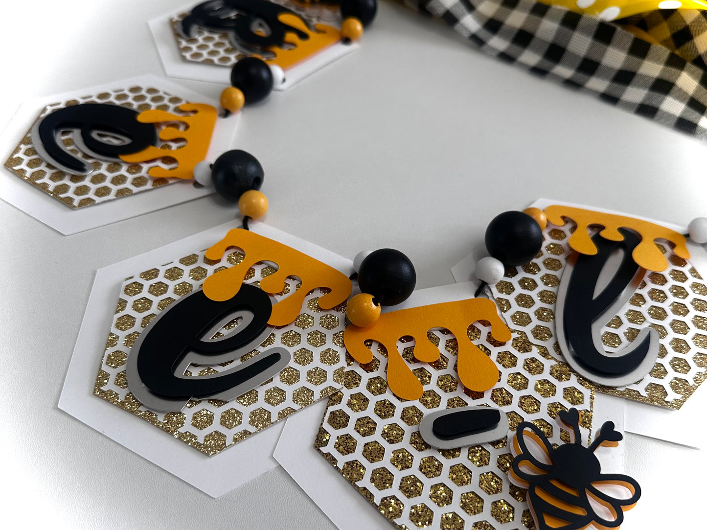 Bee-lieve handmade honeybee paper banner with glitter accents in black, yellow, and gold
Close-up of layered cardstock honeycomb details on Bee-lieve banner
Bee-lieve banner displayed as party décor with ribbon tassels and bead accents
Honeybee banner hanging in a nursery or classroom setting
Bee-lieve banner styled on a tiered tray for home décor
Full view of Bee-lieve honeybee garland banner for bee-themed celebrations